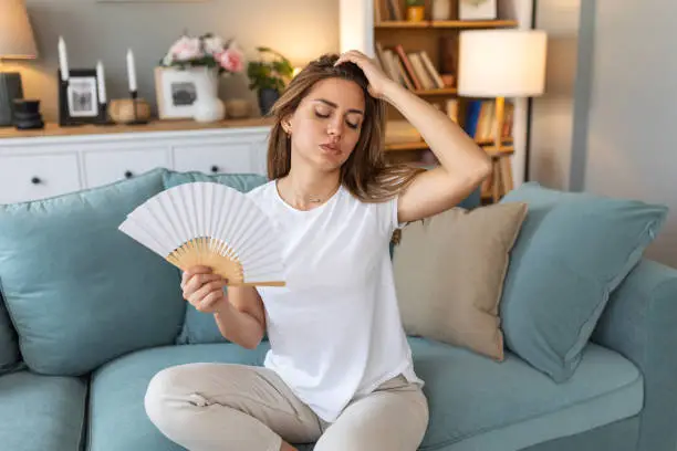 Woman puts head on sofa cushions feels sluggish due unbearable heat, waves hand fan cool herself, hot summer flat without air-conditioner climate control system concept