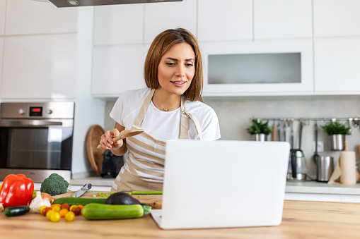 A young woman learns to cook, she watches video recipes on a laptop in the kitchen and cook a dish . Cooking at home concept