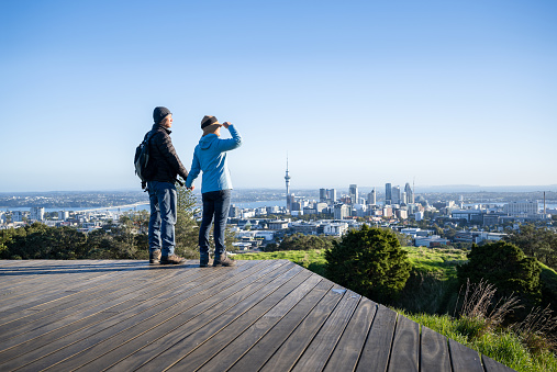 Couple standing on boardwalk at Mt Eden summit. Sky Tower and Auckland Harbour Bridge in the distance. Auckland.
