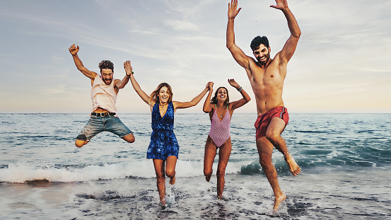 Group of young people jumping by the sea - friends having fun running by the waves - gen z people summer lifestyle concept