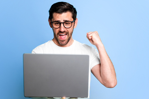 Joyful male cheers with a clenched fist while holding a laptop against a blue background