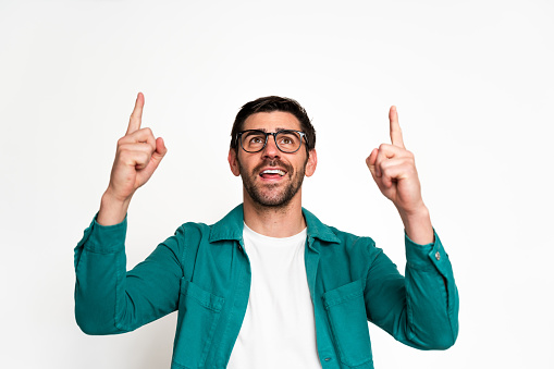 Cheerful adult male with glasses smiling and pointing upwards on a white background. Indicating joy and excitement with a positive and enthusiastic gesture while wearing casual clothing
