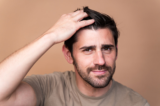 Portrait of a thoughtful young adult male with hand in hair against a beige background