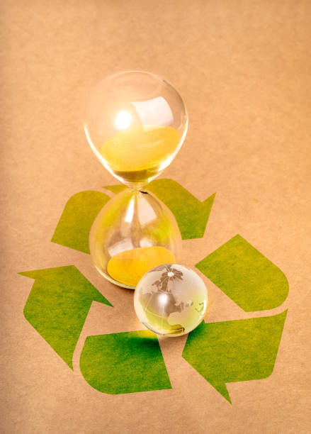 Environmental sustainability, time and responsibility, conserve resources concept. Small glass world globe near hourglass on big green recycle symbol on brown paper background, vertical style.