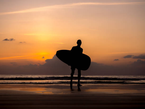 istock Man silhouette holding surfing board, walking on dark seashore. Surfer standing on sunset sky over the sea water background. Summer sand beach in evening time. dusk seascape scene. 2129503497
