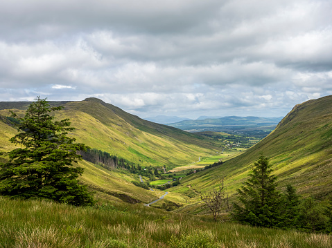 A wide view of the mountains and valleys in Ireland, with green grassy hills under grey clouds. In front is an open valley leading to distant forests and small villages. The scene captures the serene beauty of nature's landscape at Glengesh, County Donegal