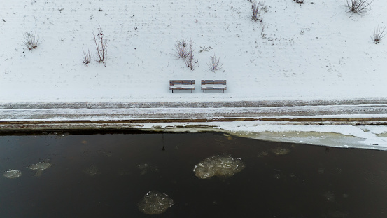 Drone photography of benches by river bank covered by snow during winter cloudy day