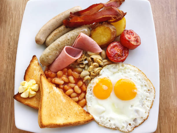 Stock photo shows a refreshing iced coffee paired with a full English fried breakfast featuring baked beans, dish of tomato, poached egg, toast, bacon, mushroom, hashbrown, and sausage. This protein-rich, hearty morning meal is perfect for brunch menus, cafe promotions