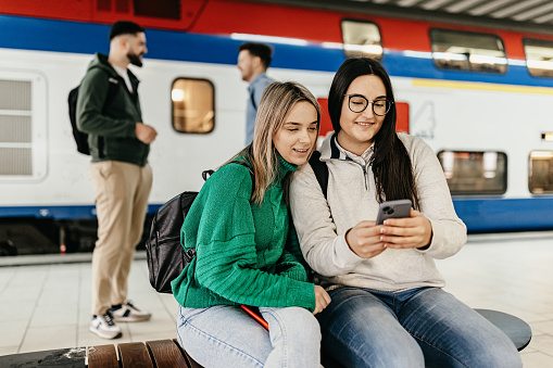 Two friends at the train station dive into their mobile phones, sharing digital moments and laughter as they wait for their train, blending the lines between virtual and real-world connections