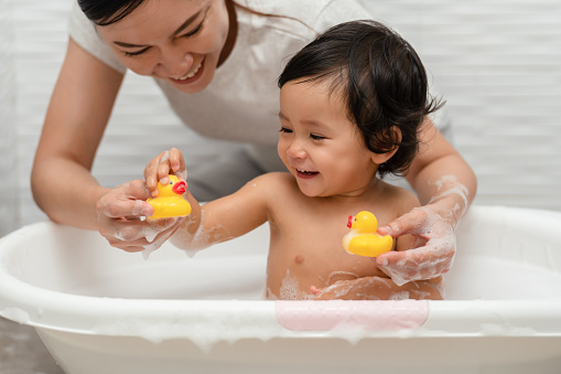mother bathing her cheerful infant baby with foam bubbles and playing rubber duck in a bathtub