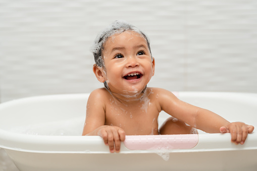 happy infant baby take a bath and playing with foam bubbles in bathtub at home