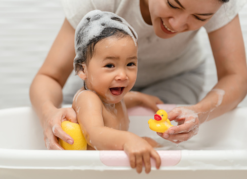mother bathing her cheerful infant baby with a soft sponge with foam bubbles in a bathtub