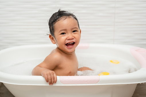 happy infant baby take a bath and playing with foam bubbles in bathtub at home