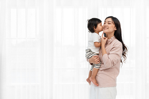happy mother being kissed by her toddler baby on white window background