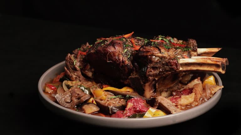 Large piece of baked or roasted meat slowly rotating on a ceramic plate on a black background. A large piece of juicy beef with bones and roasted vegetables