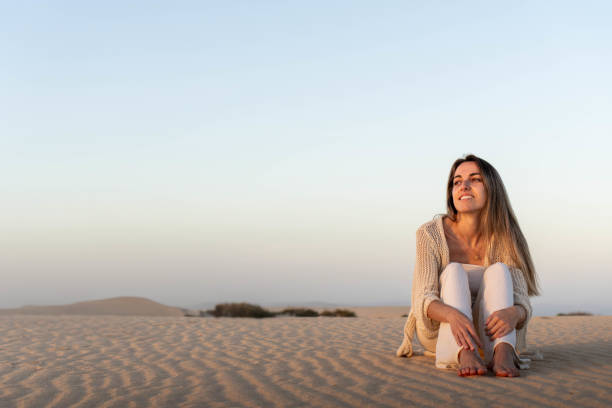 istock Happy young woman sitting on sandy dunes 2129600738