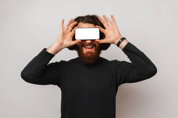Studio shot of a smiling man cover eyes with a phone with blank screen over light grey background.