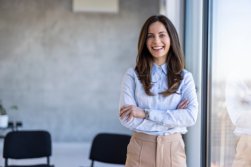 Successful businesswoman standing in creative office and looking at camera. Young woman entrepreneur in a coworking space smiling. Portrait of beautiful business woman standing in front of business team at modern agency with copy space.