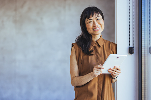 Portrait Of Female Designer Standing In Modern Office. Confident young Asian businesswoman standing smiling at the camera in a boardroom with colleagues in the background