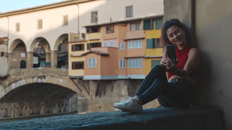 Asian girl in red T-shirt and striped blue jeans sitting on Vecchio bridge and making selfie on her smartphone. Portrait of young woman travelling to Florence during her summer vacation.