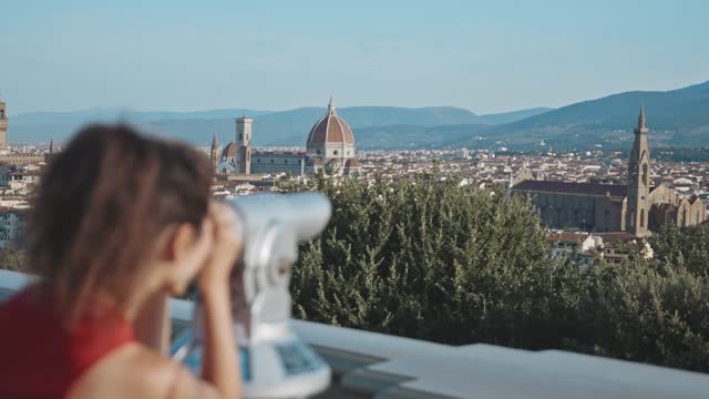 Rear view of charming young asian brunette looking through binoscope at the Cathedral of Santa Maria del Fiore. Watching the city from the viewpoint. Concept of sightseeing and travelling.