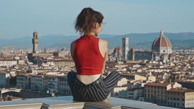 Lovely girl enjoying the beauty of the european city. Concept of travelling. Rear view of asian tourist woman sitting on the edge of viewpoint at sunset time on background of Santa Maria del Fiore.