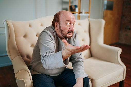 Front view aged man gesticulating with his hands trying to learn something with his eyes. Questioning look of elderly man. Confused look. Sitting indoors, at home. There are no thoughts. Lonely man.