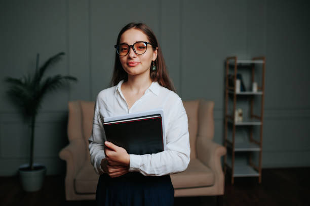 istock Young woman brunette glasses stand in office hold folder documents in hands, look in camera smile. 2129750135