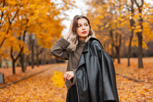 Autumn fashion portrait of beauty trendy woman model with red lips in black leather jacket with green sweater walks in golden fall park. Trendy fashion girl in fall nature
