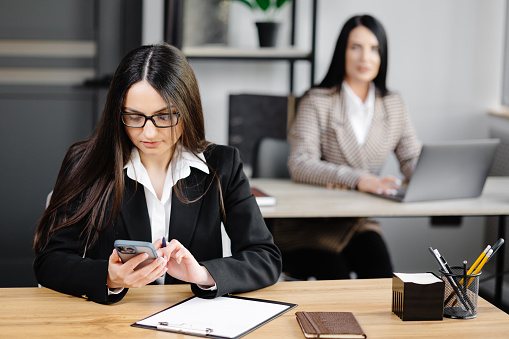Attractive young business woman wearing jacket talking on mobile phone while sitting on a desk and using notebook near her colleague at work in office.