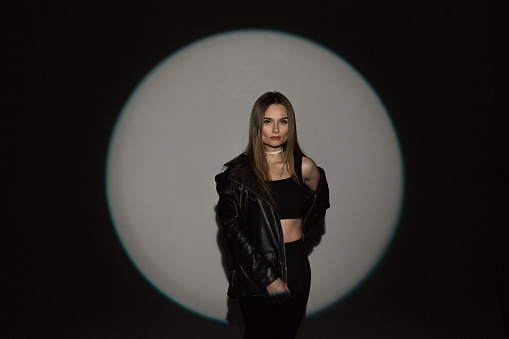 Stylish beautiful glamorous girl in fashion black rock casual clothes with top and leather jacket on a dark background with circular light at a party in the studio