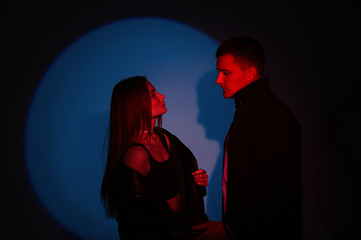 Stylish handsome man and fashionable beautiful girl in fashion clothes on dark background with creative colored red blue light