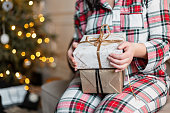 Pretty young girl in festive plaid pajamas sits on the couch and holds Christmas presents near the Christmas tree with lights on. Holidays and Christmas gifts