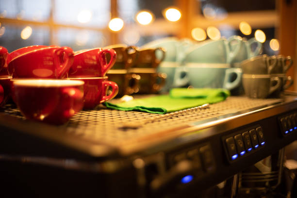 Close-up of a commercial espresso machine and coffee grinder on a terrazzo countertop in a cafe or restaurant. The setup includes stacked cups, paper cups, and coffee beans ready for brewing, highlighting modern coffee culture and professional barista equipment.