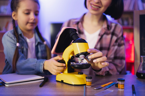 Concept of parenthood and teaching, leisure time in evening at home. Caring Caucasian mother with school daughter in casual wear working with new modern electronic microscope.