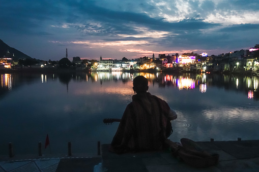 Young lonely guitarist playing on acoustic guitar in front of Pushkar Holy Lake during sunset twilight with lights reflecting on the lake over a beautiful dramatic sky
