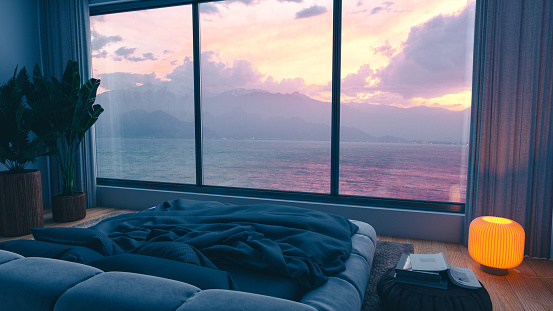 Cozy bedroom, softly lit by the warm floor lamp and evening glow, opens up to a breathtaking view of the sunset sky and sea.