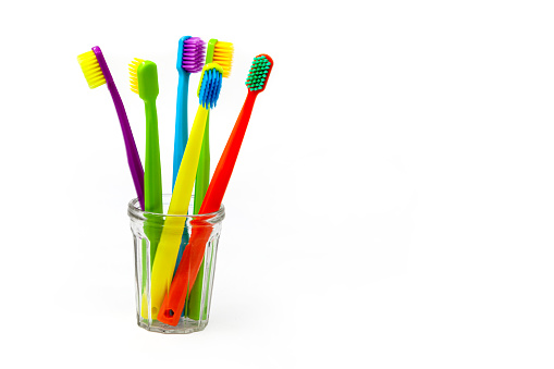 Three toothbrushes - purple, orange and yellow with different colored bristles in a clear cup on a white background. Insulated. Vertical. Photo in high quality.
