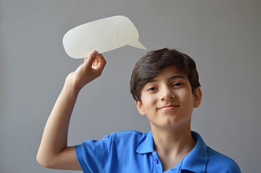 Horizontal photograph of one cute smiling 11 year old boy kid in casual blue colored t shirt holding a paper craft cut in shape of a blank empty speech bubble or call out balloon in his raised hand looking at camera with smile  over grey background