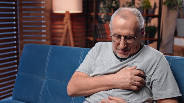 Portrait of elderly man with heartache sitting on couch in home, holding left part of chest by hand