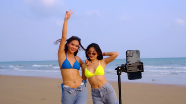 Two young Asian women with cell phone cameras stand happily dancing on the beach.