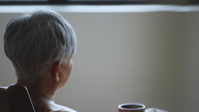 lonely asian senior woman griefing sitting in living room looking outside window with hot drink