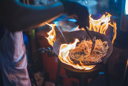 An outdoor street food chef expertly frying noodles in a flaming wok, capturing the energy of street cuisine.