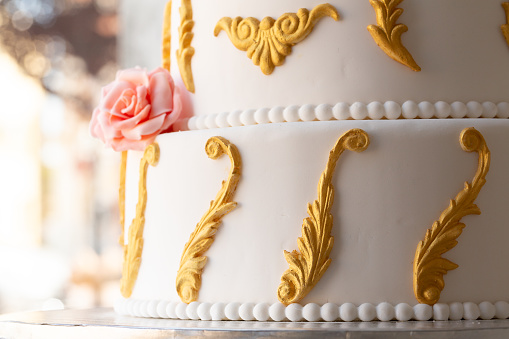an elegant wedding cake features intricate gold scrollwork and a single pink fondant rose, a true confectionery masterpiece