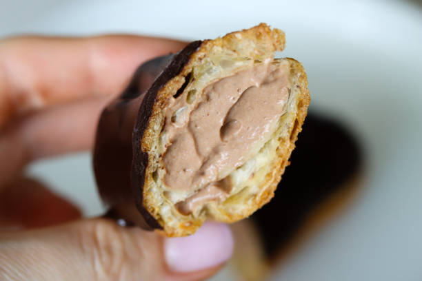 istock Close up view of halved chocolate eclair in woman's hand. Hand holding a chocolate eclair, revealing its creamy filling. Chocolate covered eclair with a rich filling, close up 2130319957