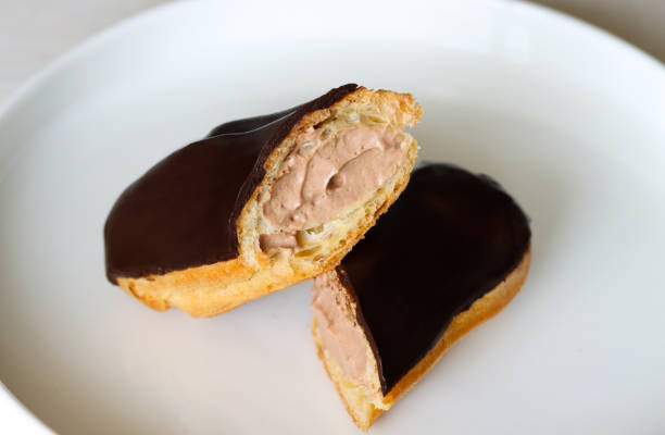 istock Chocolate eclair cut in half, showing creamy filling, on white plate, close up. Chocolate glazed eclair with rich filling on simple background. Freshly made eclair, chocolate icing and smooth filling 2130320341