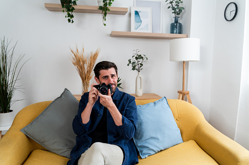 Cheerful man holding a vintage camera while sitting on a yellow sofa in a cozy living room
