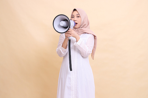 woman asian muslim wearing a hijab screams at the camera in surprise, holding a loudspeaker megaphone with both hands. Technology, broadcast and promotion concept