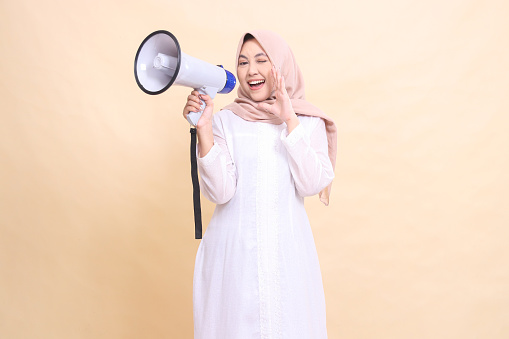 Asian Muslim woman wearing a hijab at the camera shouts cheerfully, blinking her eyes, holding a loudspeaker megaphone while her hands focus the sound. Technology, broadcast and promotion concept