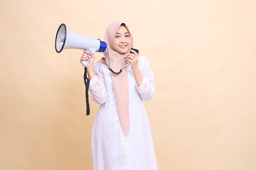 Asian Muslim woman wearing a hijab at the camera shouts, smiles, holds the speaker's megaphone to the side and holds the speaker's mic in her hand. Technology, broadcast and promotion concept
